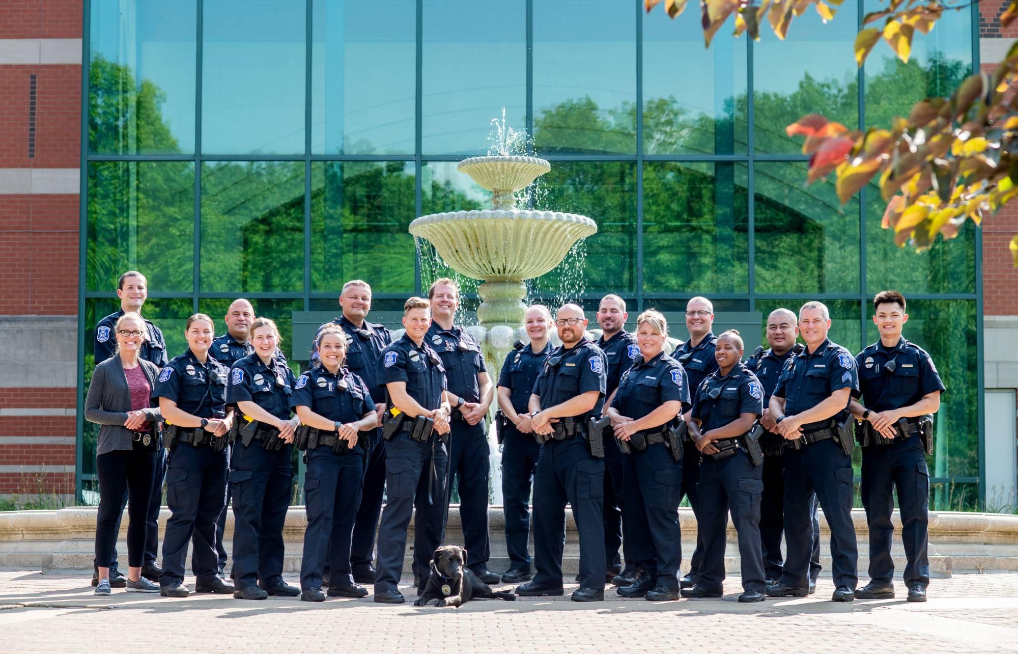 GVPD police officers in a staggered line, wearing their blue patrol uniforms, posing in front of the Student Services fountain. K9 dog Koda is laying in front. One officer, a detective is in plain clothes. The officers are smiling.